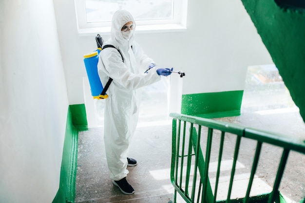 O trabalhador sanitário profissional, vestindo roupa de proteção, desinfeta um bloco de apartamentos de uma escada. Medidas de prevenção do coronavírus em áreas residenciais.