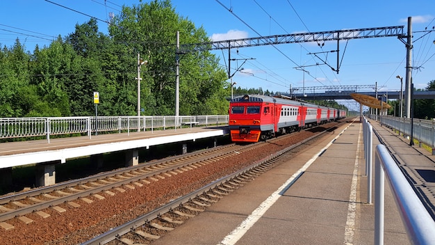 O trem suburbano chega na estação no verão no dia ensolarado. Plataforma ferroviária com trem no caminho.