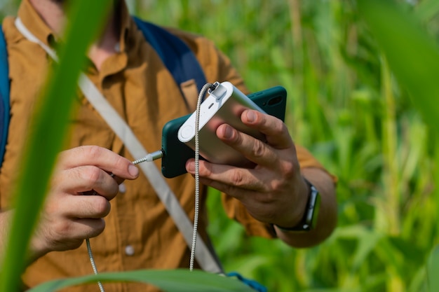 O turista tem um smartphone com um carregador portátil nas mãos. Homem com um banco de energia carrega o telefone contra o fundo do campo de milho.