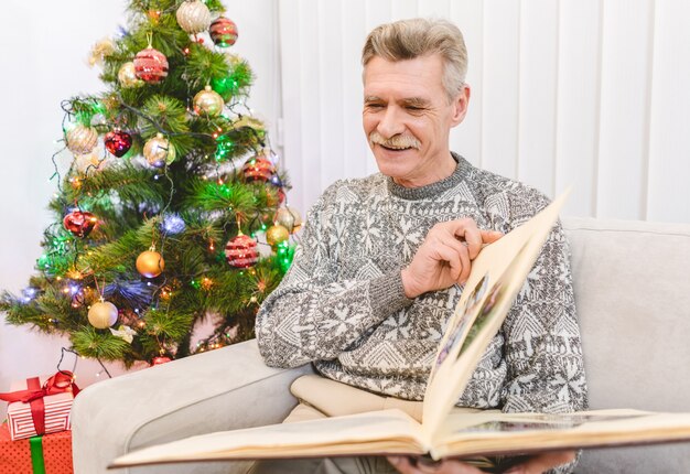 O velho feliz segurando um álbum de fotos perto da árvore de natal