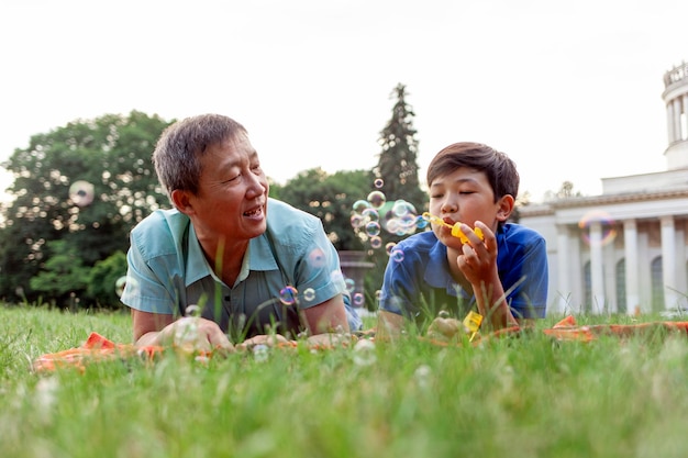 O velho pai asiático junto com o filho deitam-se na grama do parque e sopram bolhas de sabão e regozijam-se