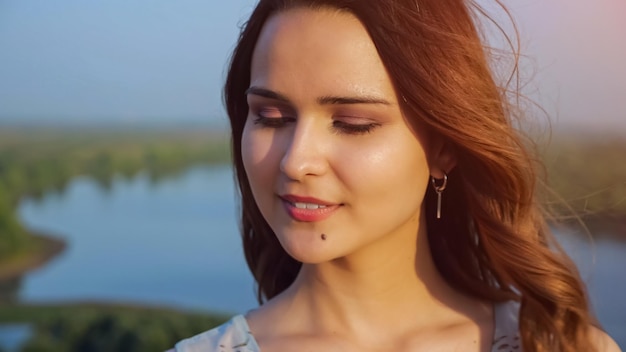 O vento brinca com mechas voadoras de uma jovem de cabelos castanhos com maquiagem leve posando para a câmera na margem do rio tranquilo na luz do sol closeup de dia de verão