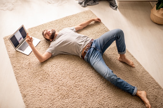 Oben Ansicht des erschöpften jungen Mannes in den Jeans, die auf Teppich im Wohnzimmer liegen und Trainer abhielten, hielt Online-Klasse