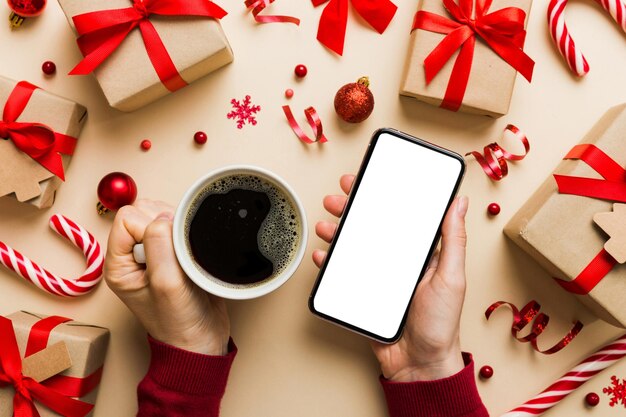 Oben Ansicht einer weiblichen Hand, die ein Smartphone mit einer heißen Tasse Kaffee hält, Weihnachtsdekoration auf farbigem Hintergrund