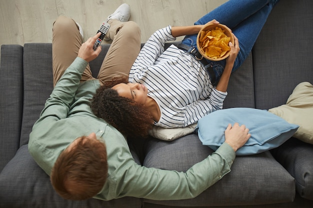 Oben Ansicht Porträt des jungen Mischlingspaares, das zu Hause fernsieht und Snacks isst, während auf bequemem Sofa liegend