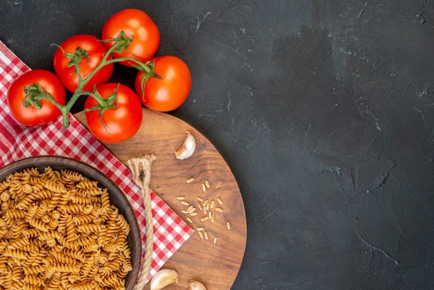 Oben Blick auf rohe Nudeln in einer braunen Schüssel auf rot abgestreiftem Handtuch Knoblauchreis auf rundem Holzbrett Tomatenseil auf der rechten Seite auf schwarzem Tisch