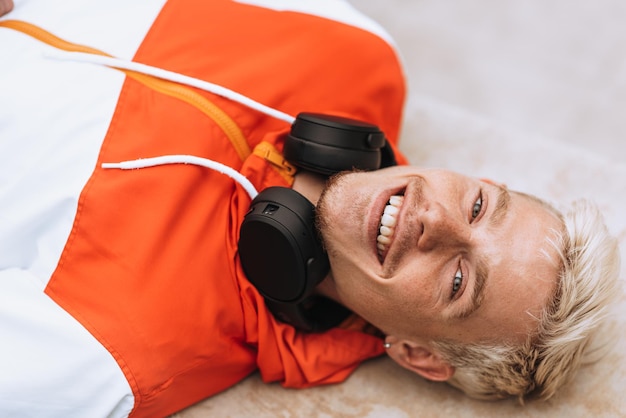 Foto oben sehen sie das bild eines gutaussehenden jungen lächelnden mannes mit glücklichem gesichtsausdruck und einem zahnigen lächeln, der mit kopfhörern am hals auf der treppe im freien in die kamera schaut lifestyle-menschen-musikkonzept