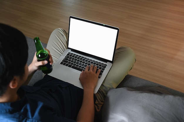 Oben sehen Sie einen lässigen Mann, der eine Flasche Bier hält und einen Laptop auf dem Sofa benutzt