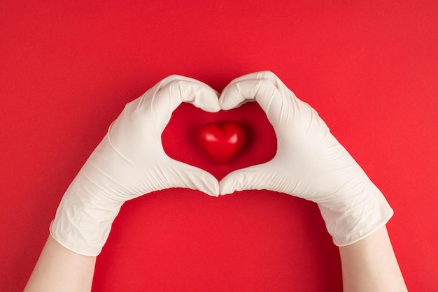 Oben über der Draufsicht Foto von weiblichen Händen in weißen Handschuhen, die Herzform und rotes Herz in der Mitte isoliert auf rotem Hintergrund machen