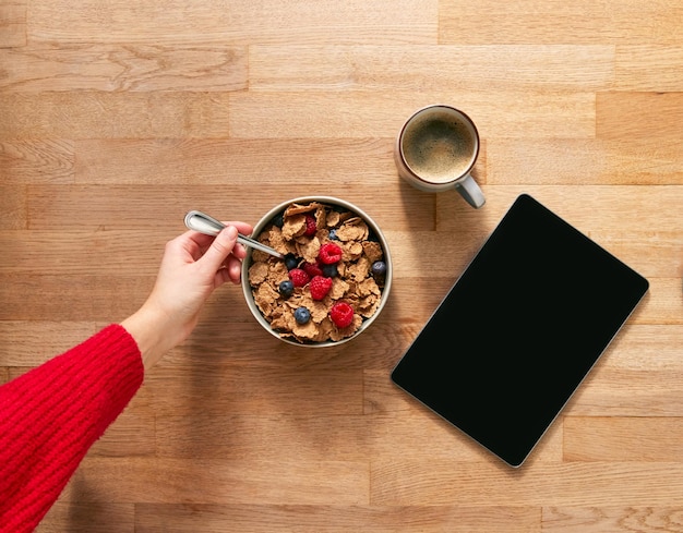 Obenliegende flache Lage der Frau mit digitalem Tablet auf dem Tisch zum Frühstück mit Müsli und Kaffee