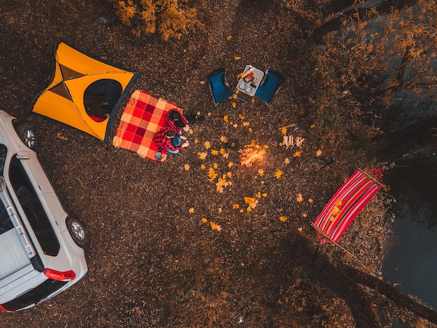 Obenliegender Blick von oben auf den Herbstcampingplatz mit Zeltautohängemattenfeuer. glückliches Paar, das sich im Freien ausruht