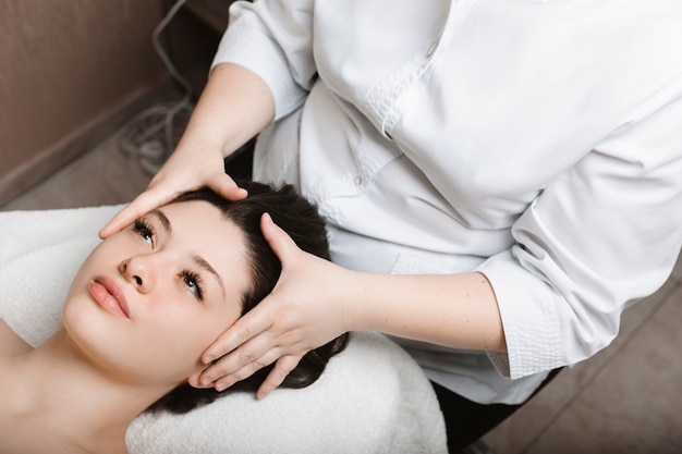 Obere Ansicht einer attraktiven Frau, die Hautpflege-Gesichtsmassage durchführt, während sie sich auf ein Spa-Bett stützt, dessen Augen in einem Wellnesscenter geöffnet sind.