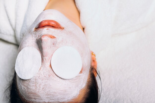 Obere Ansicht einer charmanten europäischen Frau, die auf einem Spa-Bett mit einer weißen Anti-Alter-Maske mit weißen Geweben auf ihren Augen ruht.