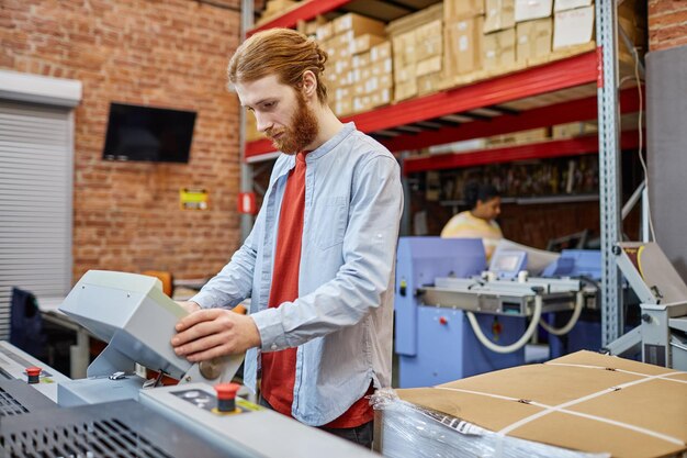 Oberkörperporträt eines bärtigen jungen Mannes, der eine Druckmaschine im Kopierraum der Werkstatt einrichtet