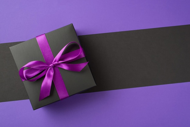 Obiges Foto einer schwarzen Geschenkbox mit violettem Band, das als Schleife gewickelt ist, isoliert auf dem violett und schwarz geteilten abstrakten Hintergrund