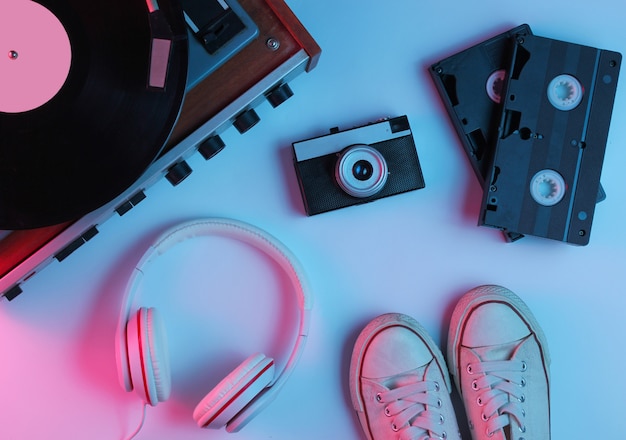 Objetos de cultura pop plana leigos retrô dos anos 80. leitor de vinil, fones de ouvido, fitas de vídeo, câmera de filme, tênis com luz gradiente de neon azul-rosa. onda retrô. vista do topo