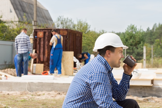 Obrero sentado tomando café relajante en un sitio en construcción mientras sus colegas continúan trabajando, vista lateral en un casco