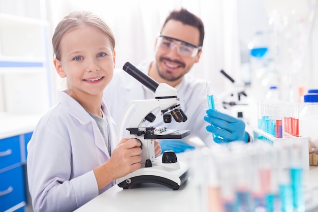 Observação emocionante. Alegre garota feliz e positiva sorrindo enquanto toca o microscópio e olha para a câmera