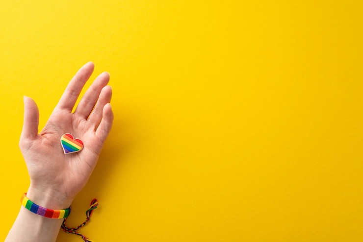 Foto observe o mês da história lgbtq com esta foto em primeira pessoa de uma mão feminina usando uma pulseira colorida de arco-íris e segurando um crachá em forma de coração em fundo amarelo com espaço para texto ou anúncio