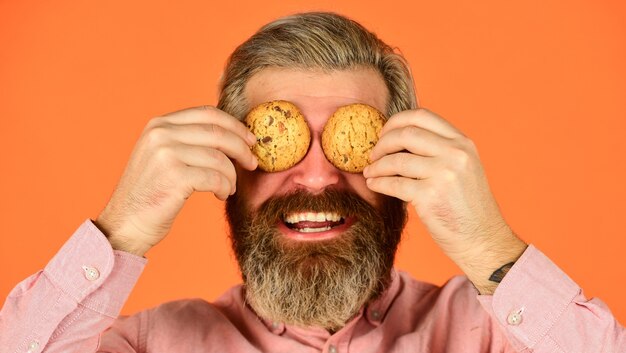 Obsesionado con la repostería. Hipster feliz comer postre de galletas. Galletas dulces. Concepto de Sweet Tooth. Cookies en su mente. Hombre barbudo sostiene galletas en los ojos. Deliciosos bocadillos deliciosos. Receta de galletas caseras.