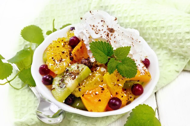 Obstsalat mit Orange, Kiwi, Cranberries und gebackenem Kürbis, Schlagsahne, bestreut mit Schokolade und Kokosnuss mit Minze in einer Schüssel auf einem Handtuch vor dem Hintergrund eines hellen Holzbretts