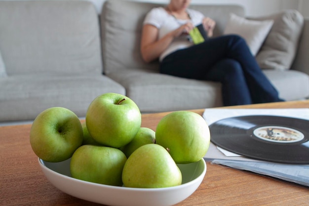 Obstschale mit grünen Äpfeln auf Holztisch und unkonzentrierte Frau, die im Hintergrund auf dem Sofa sitzt