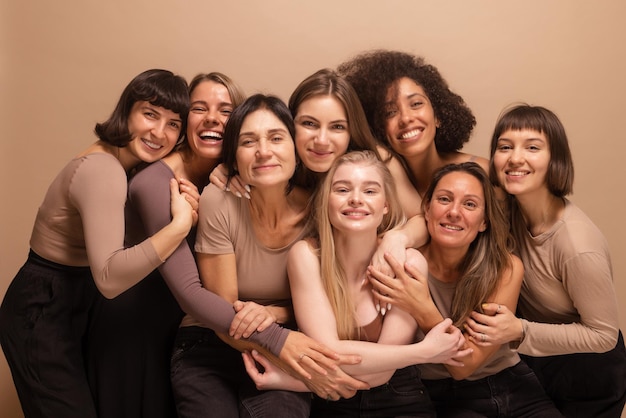 Ocho mujeres sonrientes de diferentes naciones y edades se abrazan mirando a la cámara con fondo beige