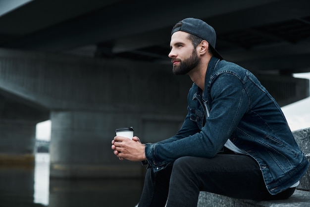 Ocio al aire libre joven elegante sentado en las escaleras en la calle de la ciudad sosteniendo la taza bebiendo caliente