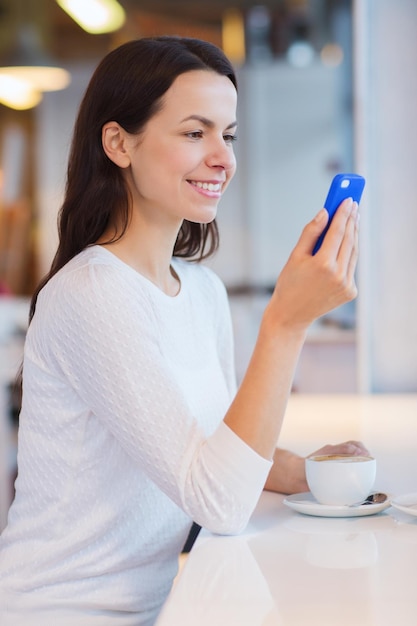 Ocio, bebidas, personas, tecnología y concepto de estilo de vida - mujer joven sonriente con teléfono inteligente tomando café en la cafetería