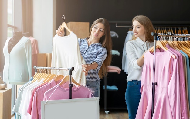 Ocio de chicas. Dos lindas chicas caucásicas eligiendo ropa, hablando y sonriendo mientras hacen compras en boutique, espacio libre