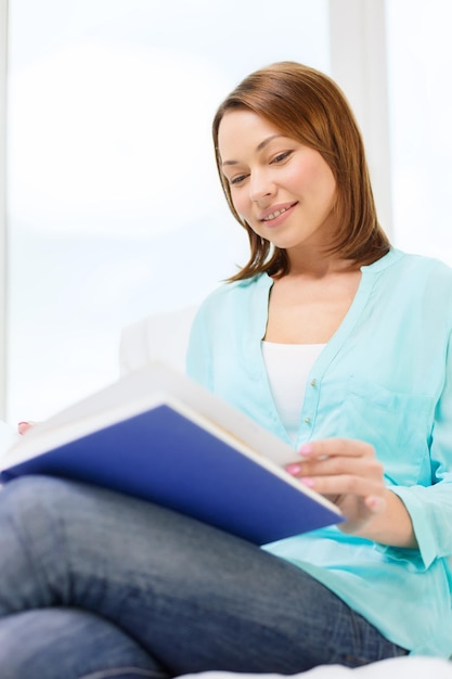 ocio y concepto de hogar - mujer sonriente leyendo un libro y sentada en el sofá en casa