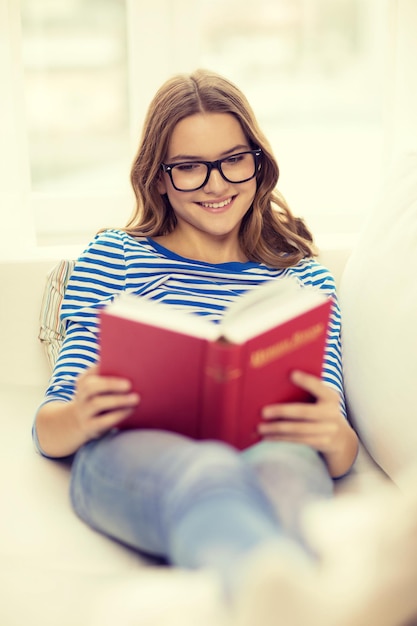 ocio, educación y concepto de hogar - adolescente sonriente con anteojos leyendo un libro y sentada en el sofá en casa