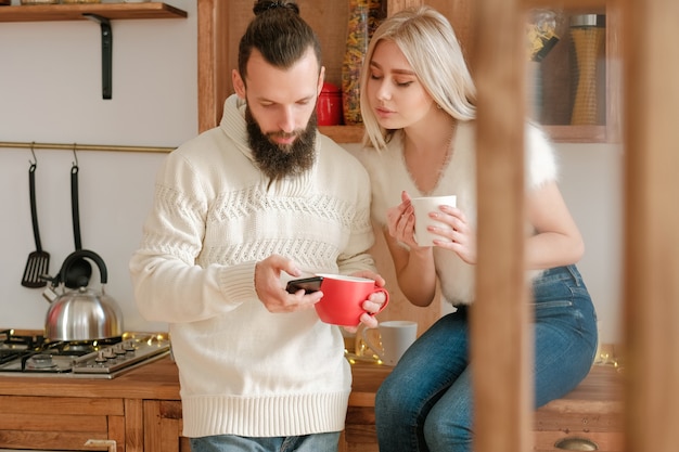 Ocio familiar por la mañana de invierno. Pareja tomando café en la cocina, usando el teléfono inteligente para enviar la invitación a la fiesta de Navidad.