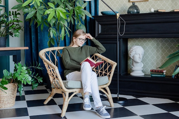 Ocio. Joven mujer rubia sentada en una silla cómoda leyendo un libro rodeado de plantas