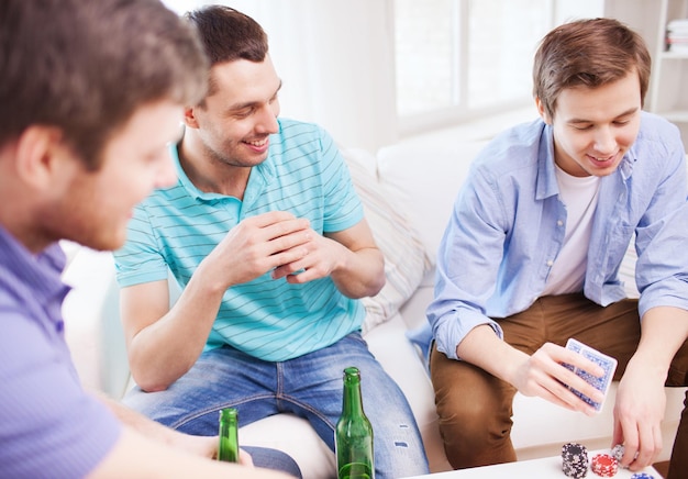ocio, juegos, amistad, juegos de azar y entretenimiento - tres amigos varones sonrientes jugando a las cartas y bebiendo cerveza en casa
