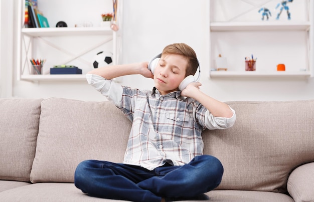 Ocio. Niño soñador escuchando música favorita en auriculares, ojos cerrados con placer. Sentado solo en el sofá en casa.