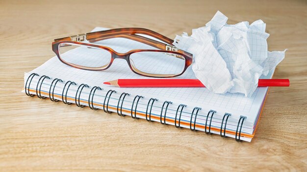 Óculos de caderno, lápis vermelho e papel amassado sobre uma superfície de madeira