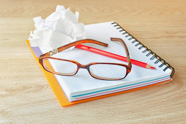 Óculos de caderno, lápis vermelho e papel amassado sobre uma superfície de madeira