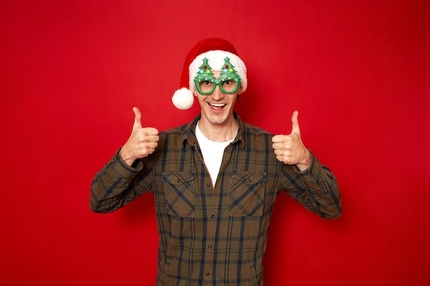 Óculos de homem animado, árvores de Natal, chapéu de Papai Noel, polegares levantados, isolados no fundo vermelho do estúdio