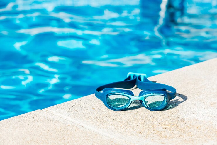 Óculos de natação azuis na calçada da piscina, no canto inferior direito, com a água azul ao longe