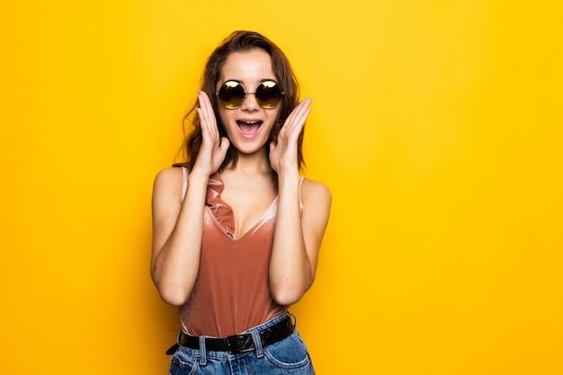 Óculos de sol de jovem olhando para longe com um sorriso surpreso isolado na parede amarela.