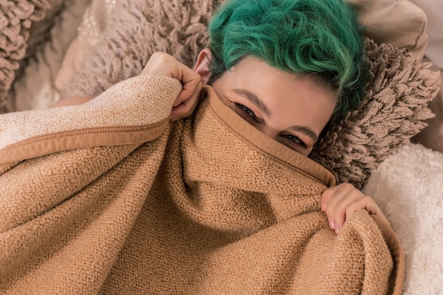 Ocultar la cara. Mujer alegre de pelo verde escondiendo su rostro detrás de cuadros en la cama por la mañana