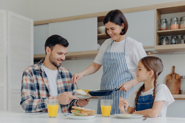 Ocupada ama de casa sostiene una sartén, da comida preparada a marido e hija, desayuna juntos