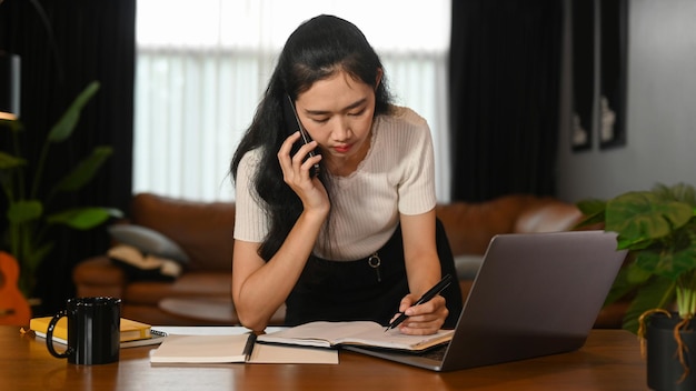 Ocupada jovem empresária conversando por telefone e fazendo anotações no notebook durante o trabalho em casa