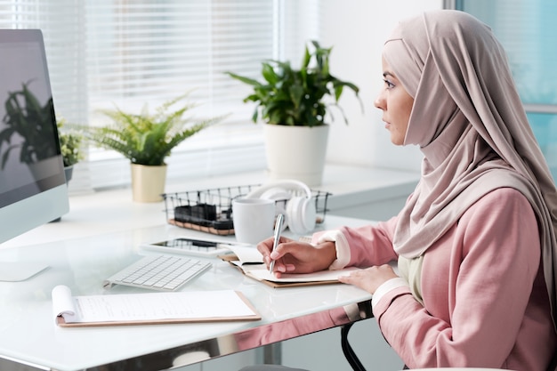 Ocupada joven musulmana en hijab sentada en la oficina moderna y tomando notas mientras trabaja con datos