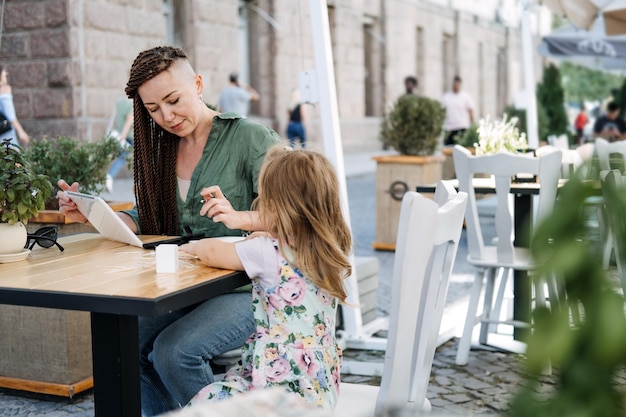 Ocupada madre exitosa y propietaria de negocios con teléfono celular y tableta desayunando con su pequeña hija en un café de la calle en el día de verano