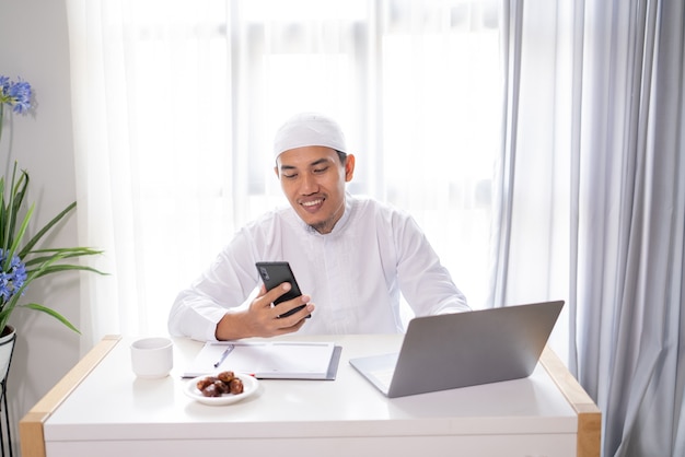 Ocupado empresario musulmán asiático usando su teléfono inteligente y computadora portátil trabajando desde casa