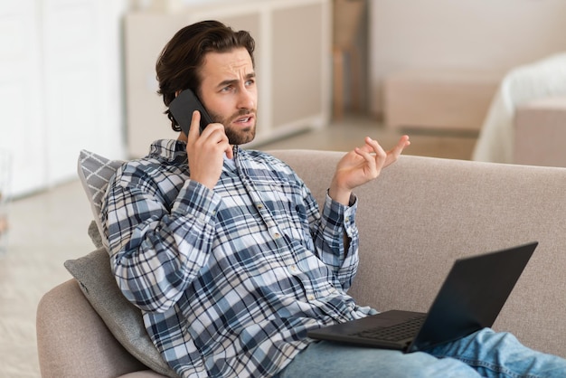 Ocupado hombre europeo milenario serio en ropa doméstica con llamadas de rastrojo en el teléfono inteligente funciona con la computadora portátil