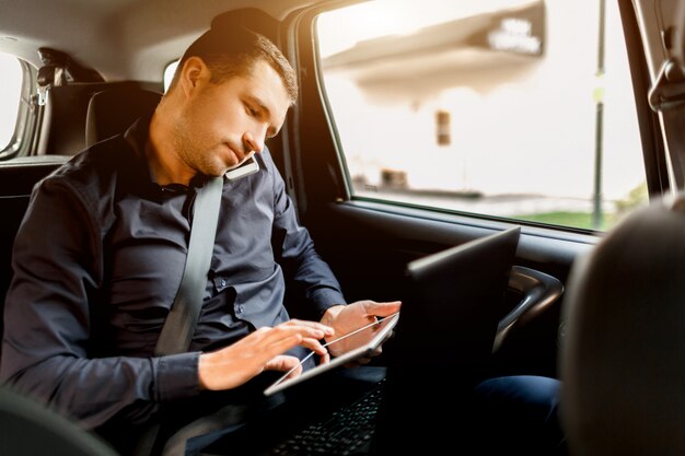 Ocupado hombre de negocios en un taxi. Concepto de multitarea El pasajero viaja en el asiento trasero y trabaja simultáneamente. Habla en un teléfono inteligente y usa una computadora portátil y una tableta.