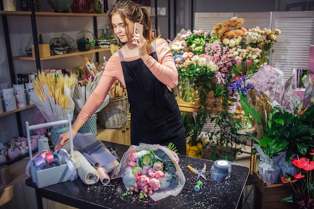 Ocupado jovem florista feminina falar no telefone. ela alcança a cesta de madeira com fitas coloridas. jovem de pé na sala cheia de flores e plantas.